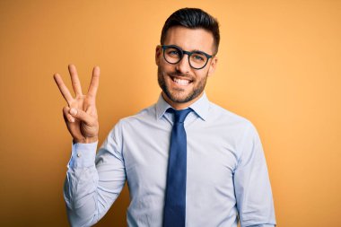 Sarı arka plan üzerinde duran kravat ve gözlük takan genç yakışıklı işadamı üç numaralı parmaklarıyla gülümseyerek kendinden emin ve mutlu bir şekilde işaret ediyor..