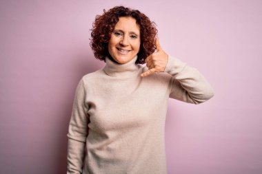 Orta yaşlı güzel kıvırcık saçlı kadın pembe arka plan üzerine balıkçı yaka kazak giyip telefonla el ve parmak hareketleri yaparak telefonda konuşur gibi gülümsüyor. İletişim kavramları.