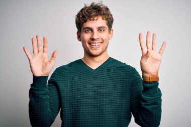 Sarışın, kıvırcık saçlı, beyaz arka planda yeşil süveter giyen ve dokuz numaralı parmaklarıyla işaret eden genç, kendinden emin ve mutlu bir adam..