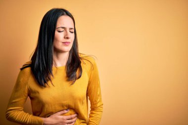 Young brunette woman with blue eyes wearing casual sweater over yellow background with hand on stomach because indigestion, painful illness feeling unwell. Ache concept. clipart