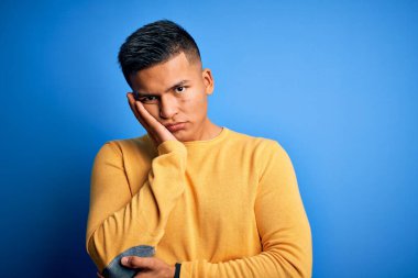 Sarı kazak giyen genç, yakışıklı, latin bir adam. Mavi arka planda, kollarını kavuşturarak depresyondan sıkılmış ve yorgun görünüyor..