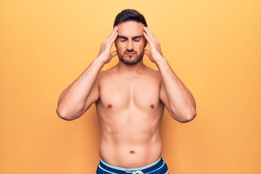 Sakallı, kolsuz tişörtlü genç ve yakışıklı bir adam sarı arka planda duruyor. Ağrı ve migren yüzünden baş ağrısı ve stresten muzdarip. Ellerini başının üstüne koy..