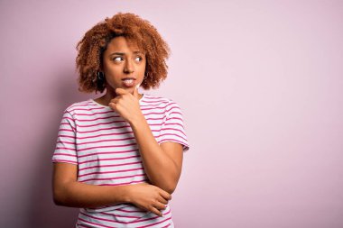 Kıvırcık saçlı, sıradan çizgili tişörtlü, eli çenesinde soru soran Afro-Amerikan bir kadın. Düşünceli bir ifade. Düşünceli bir yüzle gülümsüyor. Şüphe kavramı.