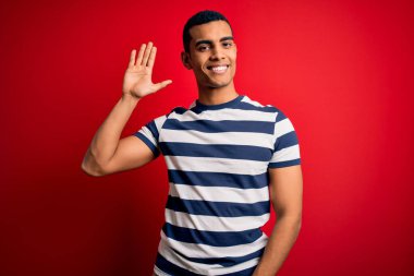 Yakışıklı Afro-Amerikalı sıradan çizgili tişört giyen kırmızı arka planda duran mutlu ve güler yüzlü hoş geldin jesti yapan