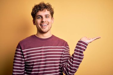 Sarışın, kıvırcık saçlı, sıradan çizgili süveter giyen neşeli bir şekilde gülümseyen ve avuç içi kameraya bakan genç bir adam..