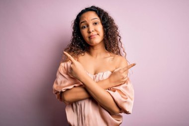 Kıvırcık saçlı, rahat tişörtlü, pembe arka planda duran, iki tarafı da parmaklarıyla gösteren, farklı yönlü, güzel bir kadın.