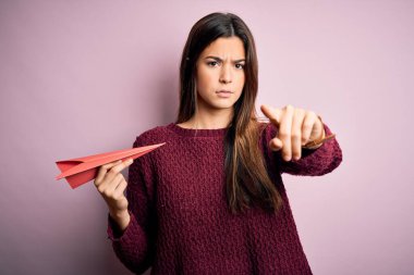 Elinde kağıttan bir uçak tutan genç ve güzel bir kız pembe arka plan üzerinde duruyor. Kamerayı işaret ediyor ve sen, el işareti, ön taraftan pozitif ve kendinden emin bir jest.