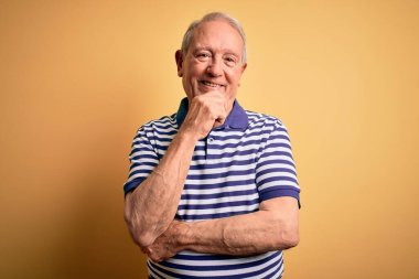 Gri saçlı, donanma çizgili tişört giyen, sarı arka planda duran ve kameraya güvenen kolları çapraz ve çenesi havada gülümseyen yaşlı bir adam. Olumlu düşünüyorum..