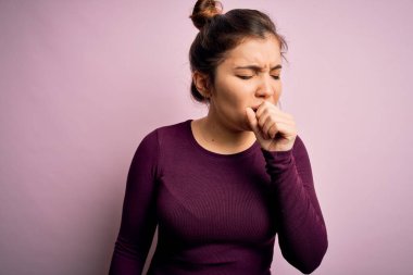 Güzel genç bir kadın pembe izole edilmiş arka planda günlük çörek saç stili takıyor kendini iyi hissetmiyor ve soğuk algınlığı veya bronşitin belirtisi olarak öksürüyor. Sağlık hizmeti kavramı.