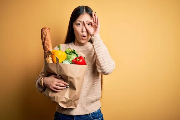 年轻的亚洲女人拿着装有新鲜健康食品的纸袋 在黄色孤立的背景上做着一个很好的姿势 用惊讶的脸 用手指看着 难以置信的表情 — 图库照片