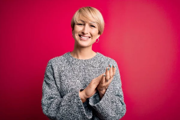 Modern kısa saçlı genç bir kadın pembe arka planda sıradan bir süveter giyiyor ve mutlu ve neşeli elleri alkışlıyor.