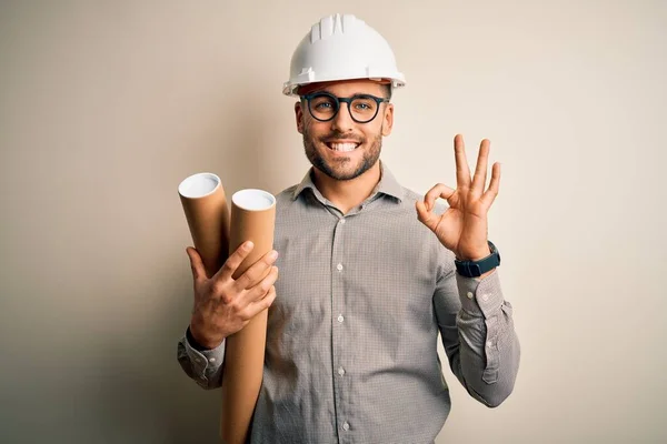 若い建築家の男は 指でOkサインを行う孤立した背景にプロジェクトの紙計画を保持請負業者ヘルメットを身に着けている 優れたシンボル — ストック写真