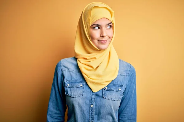 若いです美しいです女の子身に着けていますムスリムヒジャーブ立ちます上の隔離された黄色の背景笑顔見ますへ側と離れて考え — ストック写真