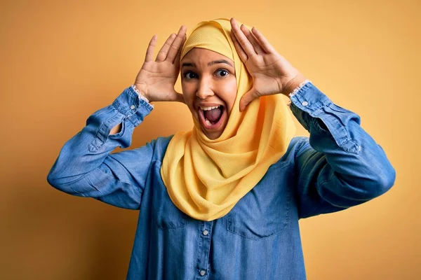 黄色の背景にアラブの伝統的なヒジャーブを身に着けている巻き毛を持つ若い美しい女性明るい顔を示す手で竹を覗かせる笑みを浮かべて 驚きと興奮した — ストック写真