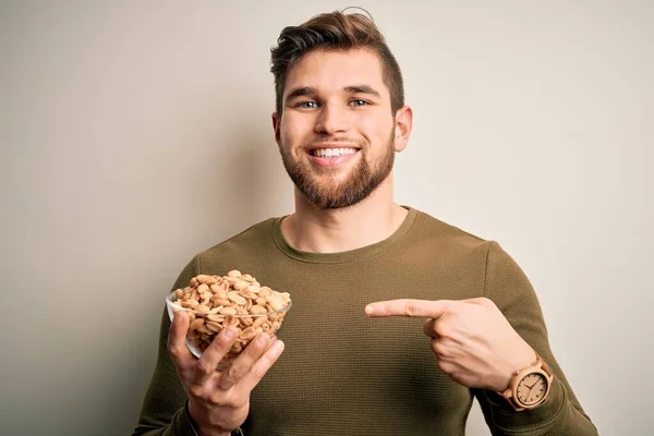 Young blond man with beard and blue eyes holding bowl with healthy salty peanuts very happy pointing with hand and finger