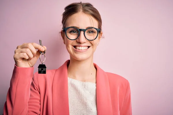 Young beautiful redhead house agent woman holding home key over pink bakcground with a happy face standing and smiling with a confident smile showing teeth