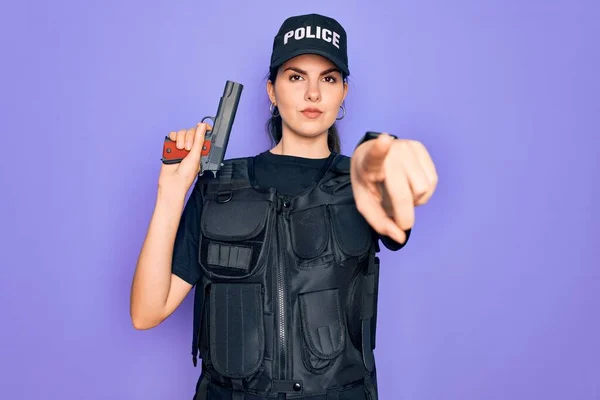 Young police woman wearing security bulletproof vest uniform and holding gun pointing with finger to the camera and to you, hand sign, positive and confident gesture from the front