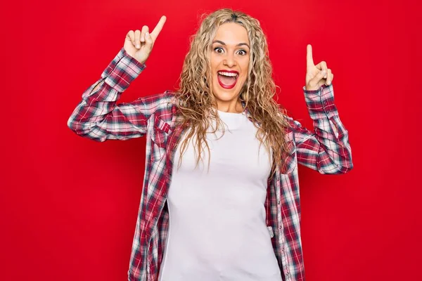 Genç ve güzel sarışın kadın, gündelik gömleğini giyip kırmızı arka planda dikiliyor. Şaşırmış, şaşırmış ve parmaklarıyla işaret ediyor ve kollarını kaldırıyor..
