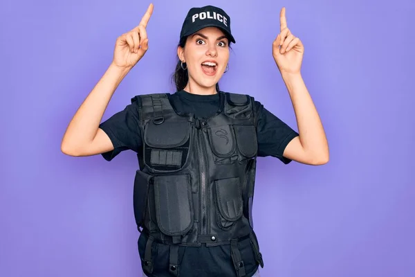 Young police woman wearing security bulletproof vest uniform over purple background smiling amazed and surprised and pointing up with fingers and raised arms.