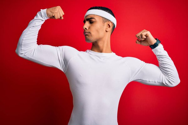 Молодой красивый африканский спортсмен, одетый в спортивную форму на красном фоне, показывает мышцы рук, улыбающиеся с гордостью. Фитнес-концепция
.