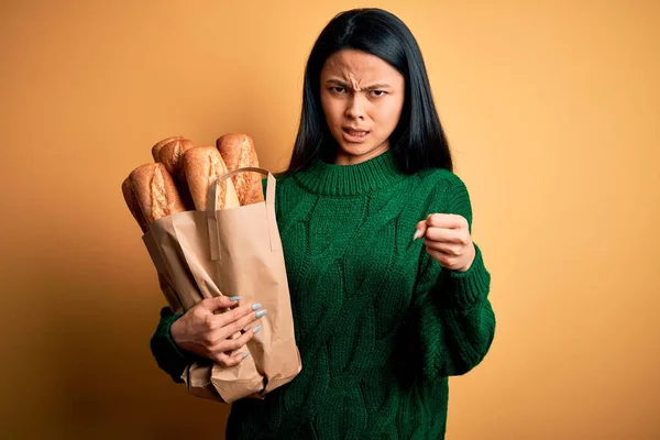 年轻美丽的中国女人拿着装有面包的纸袋 披着孤立的黄色背景 怒气冲冲地大叫 疯狂地高喊 怒气冲冲 — 图库照片