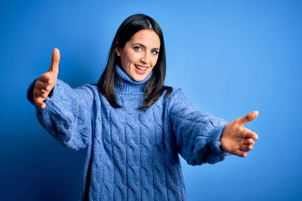 Young brunette woman with blue eyes wearing casual turtleneck sweater looking at the camera smiling with open arms for hug. Cheerful expression embracing happiness.