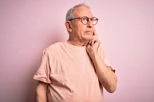 Grey haired senior man wearing glasses standing over pink isolated background with hand on chin thinking about question, pensive expression. Smiling with thoughtful face. Doubt concept.