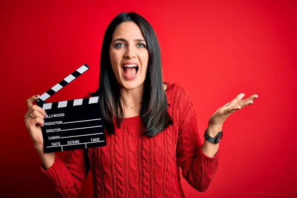 Young director woman with blue eyes making movie holding clapboard over red background very happy and excited, winner expression celebrating victory screaming with big smile and raised hands