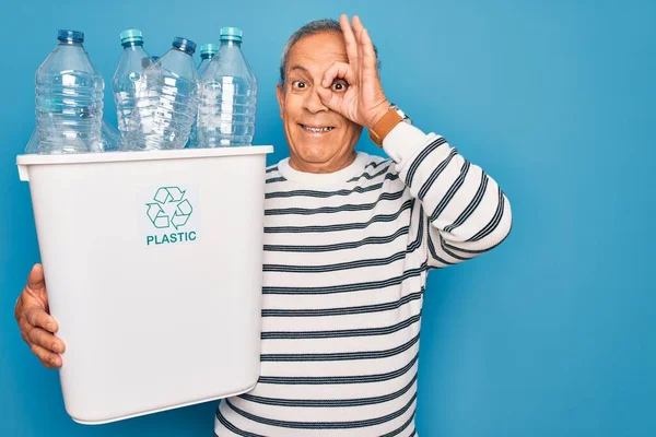 シニア男リサイクル保持ゴミ缶プラスチックボトルでリサイクル上の青の背景に幸せな顔を笑顔で手でOkサインを行う指を通して見て — ストック写真