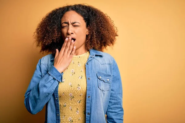 Young african american woman with afro hair wearing casual denim shirt over yellow background bored yawning tired covering mouth with hand. Restless and sleepiness.