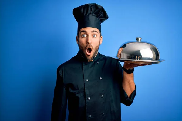 年轻的厨师 留着胡子 身穿制服 拿着圆顶蓝色背景的盘子 带着惊讶的表情 害怕而兴奋 — 图库照片