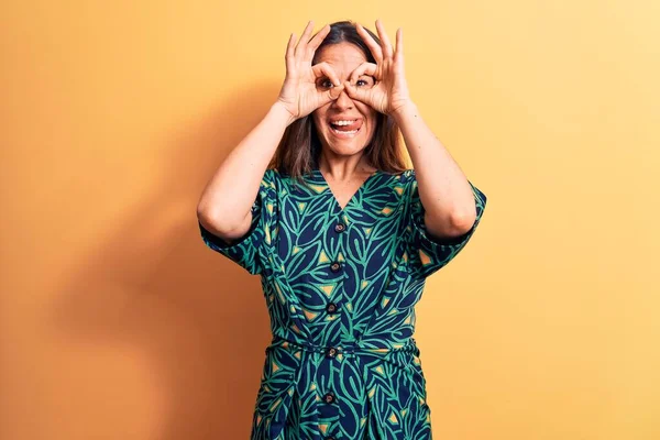 Sarı arka planda dikilen sıradan çiçekli bir elbise giyen genç, güzel esmer kadın dürbün gibi jest yapıyor, gözleri parmaklarından bakıyor. Çılgın ifade.