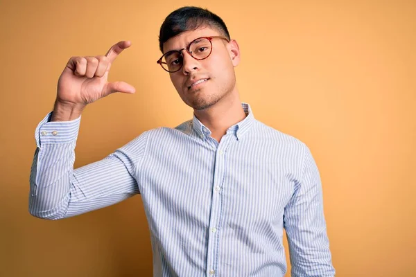 Young handsome hispanic business man wearing nerd glasses over yellow background smiling and confident gesturing with hand doing small size sign with fingers looking and the camera. Measure concept.