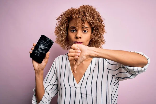 Young african american curly woman holding broken smartphone showing craked screen with angry face, negative sign showing dislike with thumbs down, rejection concept