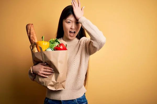 年轻的亚洲女人拿着装有新鲜健康食品的纸袋 在黄色孤立的背景上 手头上的错误让人吃惊 记住错误 记性差 — 图库照片