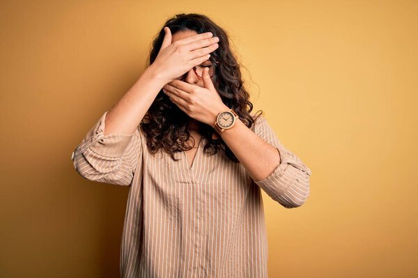 Beautiful woman with curly hair wearing striped shirt and glasses over yellow background Covering eyes and mouth with hands, surprised and shocked. Hiding emotion