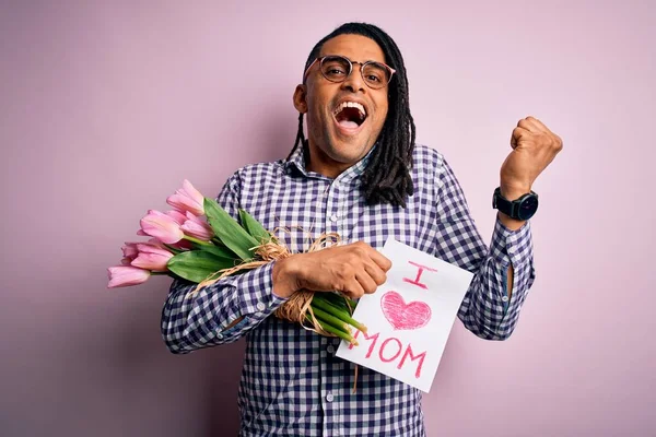 African american man with dreadlocks holding love mom message and tulips on mothers day screaming proud and celebrating victory and success very excited, cheering emotion — Stock Image African American Man Dreadlocks Holding Love Mom Message Tulips Mothers Stock Photo