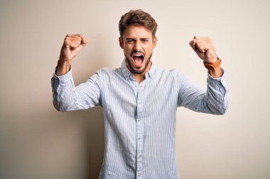 Sakallı, çizgili tişörtlü genç bir adam beyaz arka planda kızgın ve kızgın duruyor. Öfkeyle bağırırken yumruklarını kaldırıyor ve sinirleniyor. Öfke ve agresif kavram.