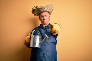 Sarı arka plan üzerinde önlük ve şapka tutan orta yaşlı çiftçi kamerayı işaret ediyor ve sen, el işareti, ön taraftan pozitif ve kendinden emin bir hareket.