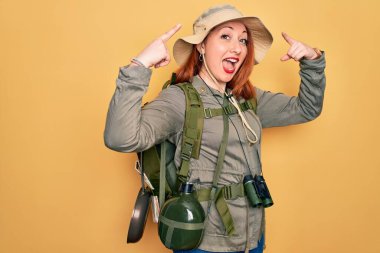 Sırt çantası ve sarı arka planda şapka takan genç kızıl saçlı kadın iki eliyle kafasını gösteren gülümseme, harika bir fikir ya da düşünce, iyi bir hafıza.