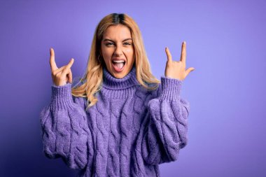 Güzel sarışın kadın, mor arka plan üzerine balıkçı yaka kazak giyip çılgın bir ifadeyle elleri havada rock sembolü yaparak bağırıyor. Müzik yıldızı. Ağır müzik konsepti.