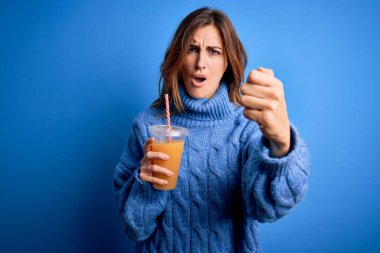Genç, güzel esmer kadın sağlıklı portakal suyunu mavi arka planda içerken sinirlenmiş ve sinirlenmiş öfkeyle bağırıyor, çıldırmış ve el kaldırarak bağırıyor.