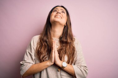 Genç, güzel, esmer bir kadın pembe arka planda sıradan bir süveter giyiyor. Elleri bağlı yalvarıyor ve dua ediyor. Yüzünde umut ifadesi var. Yalvarıyorum..