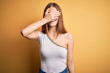Genç, güzel, kızıl saçlı bir kadın izole edilmiş sarı arka planda günlük tişört giyiyor. Gülümsüyor ve yüzünde şaşkınlıktan gözleri kapayarak gülüyor. Kör kavram.