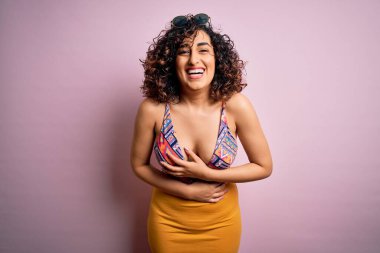 Genç, güzel bir Arap kadın, mayo ve pembe arka planda güneş gözlüğü takmış, gülümsüyor ve kahkahalarla gülüyor..