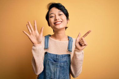 Genç, güzel Asyalı kız izole edilmiş sarı arka planda günlük kot pantolon giyiyor ve yedinci parmaklarını gösterirken kendinden emin ve mutlu bir şekilde gülümsüyor..