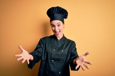 Young beautiful chef woman wearing cooker uniform and hat standing over yellow background smiling cheerful offering hands giving assistance and acceptance. clipart