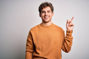 Sarışın, kıvırcık saçlı, beyaz arka planda sıradan bir süveter giyen, yüzünde kocaman bir gülümseme olan, eli kameraya bakan genç ve yakışıklı bir adam..