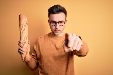 Genç, beyaz, gözlüklü bir adam elinde taze ekmek ekmeği ile kamerayı işaret ediyor ve sen, el işareti, ön taraftan pozitif ve kendinden emin bir jest.