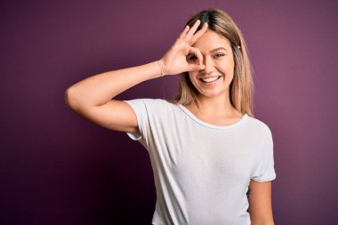Genç, güzel, sarışın bir kadın mor izole edilmiş arka plan üzerine günlük beyaz tişört giyiyor. El hareketleriyle gülümsüyor, gözleriyle mutlu yüzle parmaklarının arasından bakıyor..